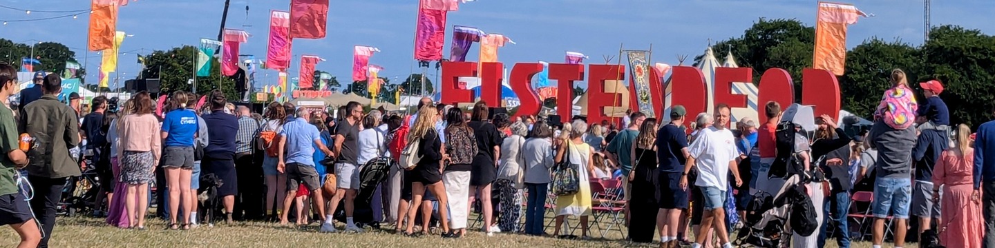 Ymwelwyr yn mwynhau cerdded ar faes yr Eisteddfod gyda'r haul yn tywynnu dros lythrennau enwog yr Eisteddfod.